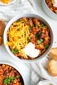 A bowl of hearty beef chili topped with shredded cheddar cheese, sour cream, and fresh cilantro, served with toasted baguette slices.