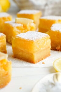 Close-up of a thick lemon bar with a buttery shortbread crust and vibrant lemon filling, dusted with powdered sugar and surrounded by lemon wedges.