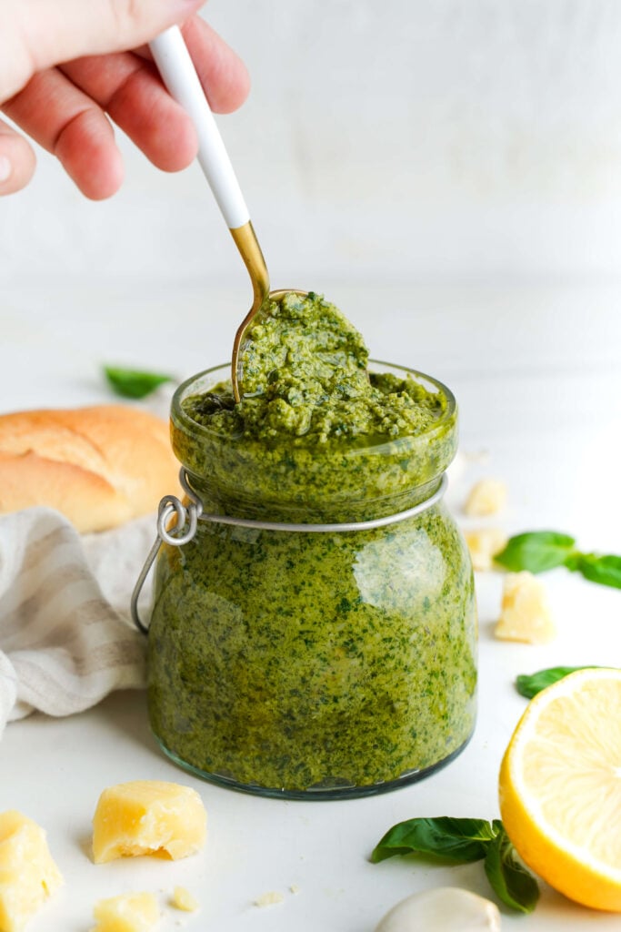 Homemade pesto in a glass jar with a spoon, surrounded by basil, Parmesan, lemon, and bread.