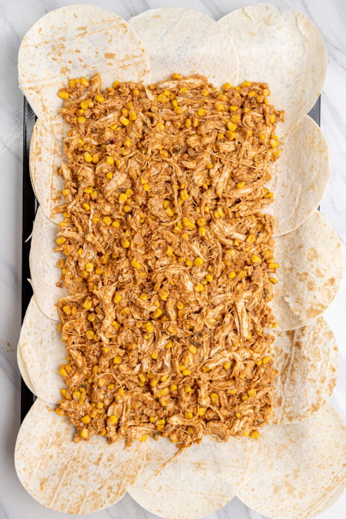 Sheet pan chicken quesadilla filling with shredded chicken and corn spread evenly over overlapping flour tortillas on a baking sheet