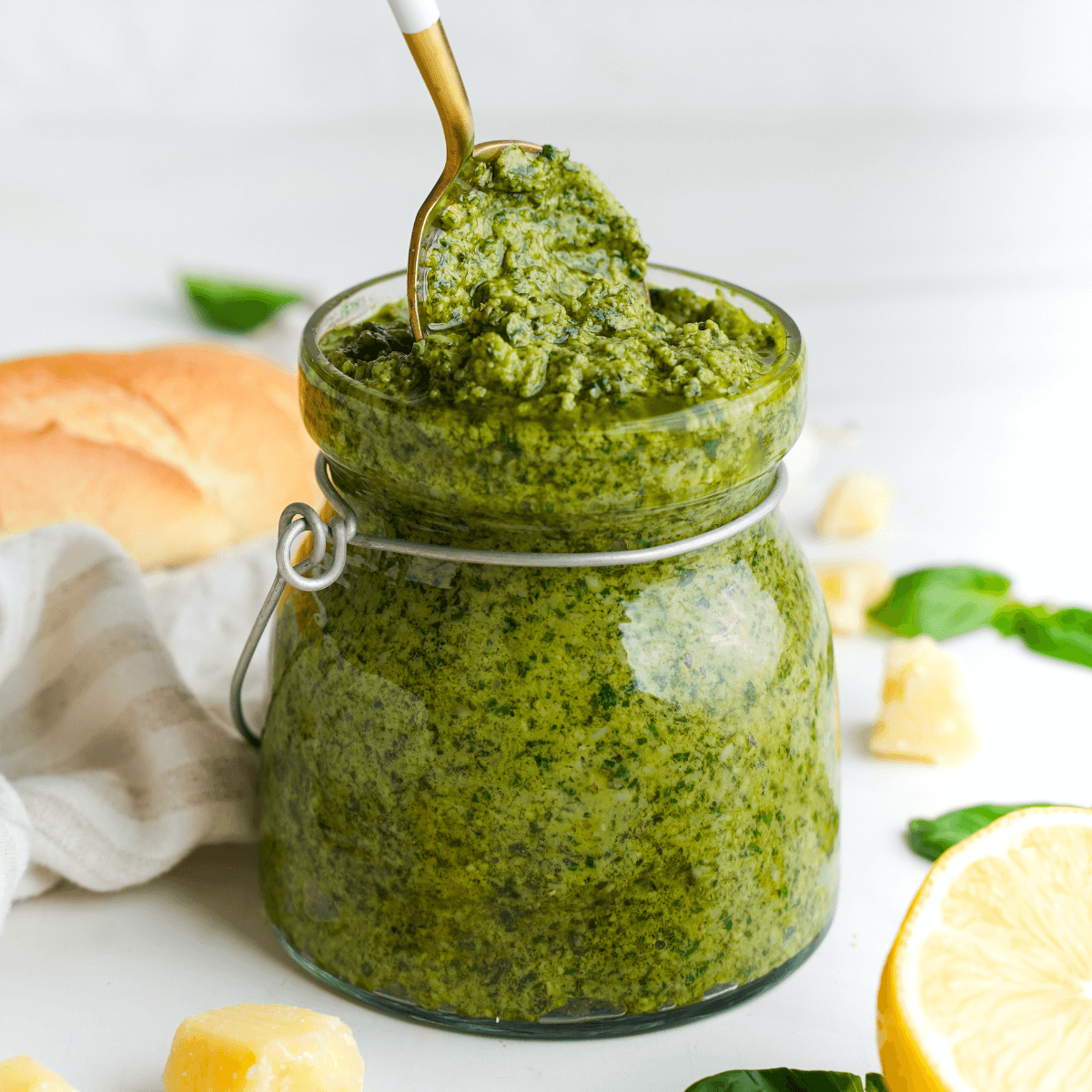 Homemade pesto in a glass jar with a spoon, surrounded by basil, Parmesan, lemon, and bread.