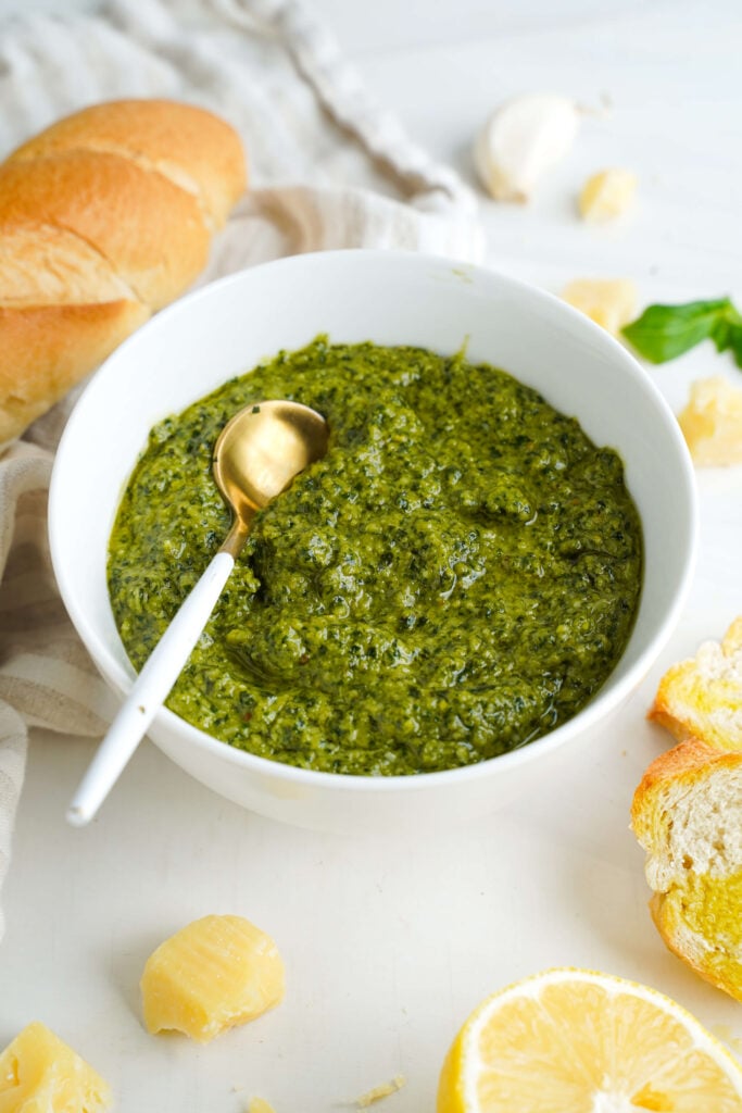 Basil pesto served in a white bowl with a gold spoon, surrounded by Parmesan, lemon, and crusty bread
