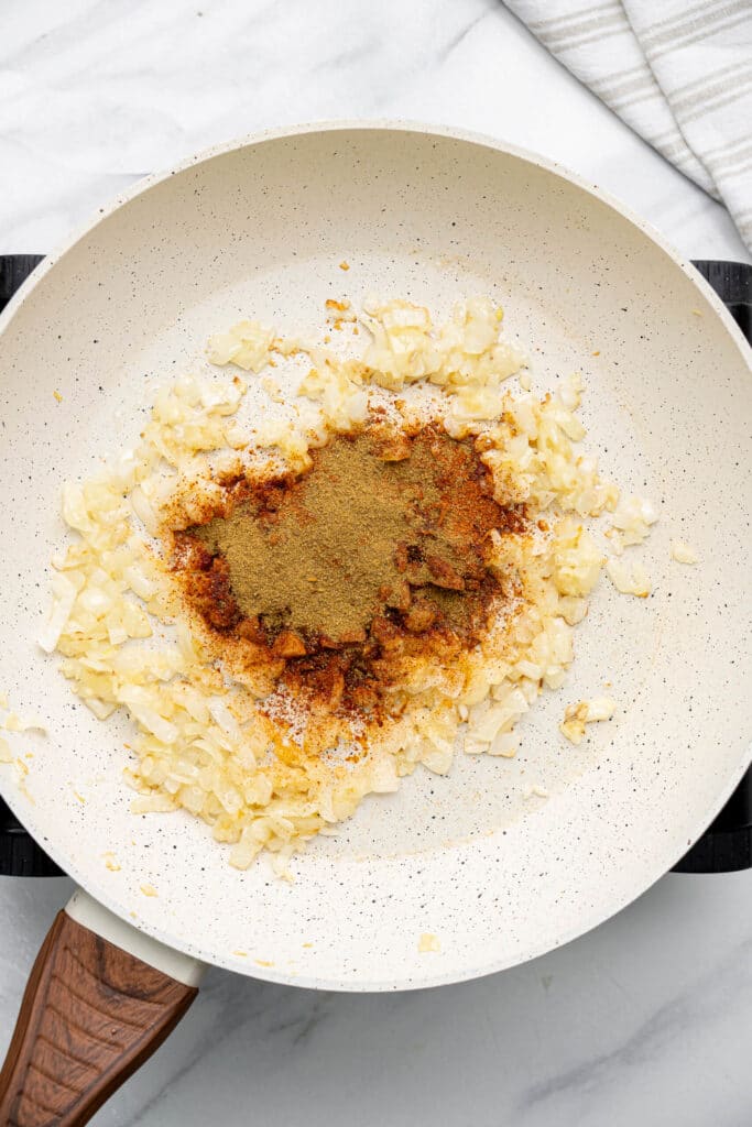 Chili powder, cumin, and other spices added to sautรฉed onions in a skillet