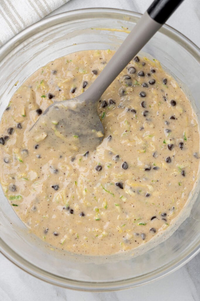 Zucchini muffin batter mixed with chocolate chips in a glass bowl with a grey spatula, ready to be scooped into muffin liners.