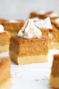 A close-up of a pumpkin pie bar with a thick pumpkin filling on a shortbread crust, topped with whipped cream and a dusting of cinnamon.