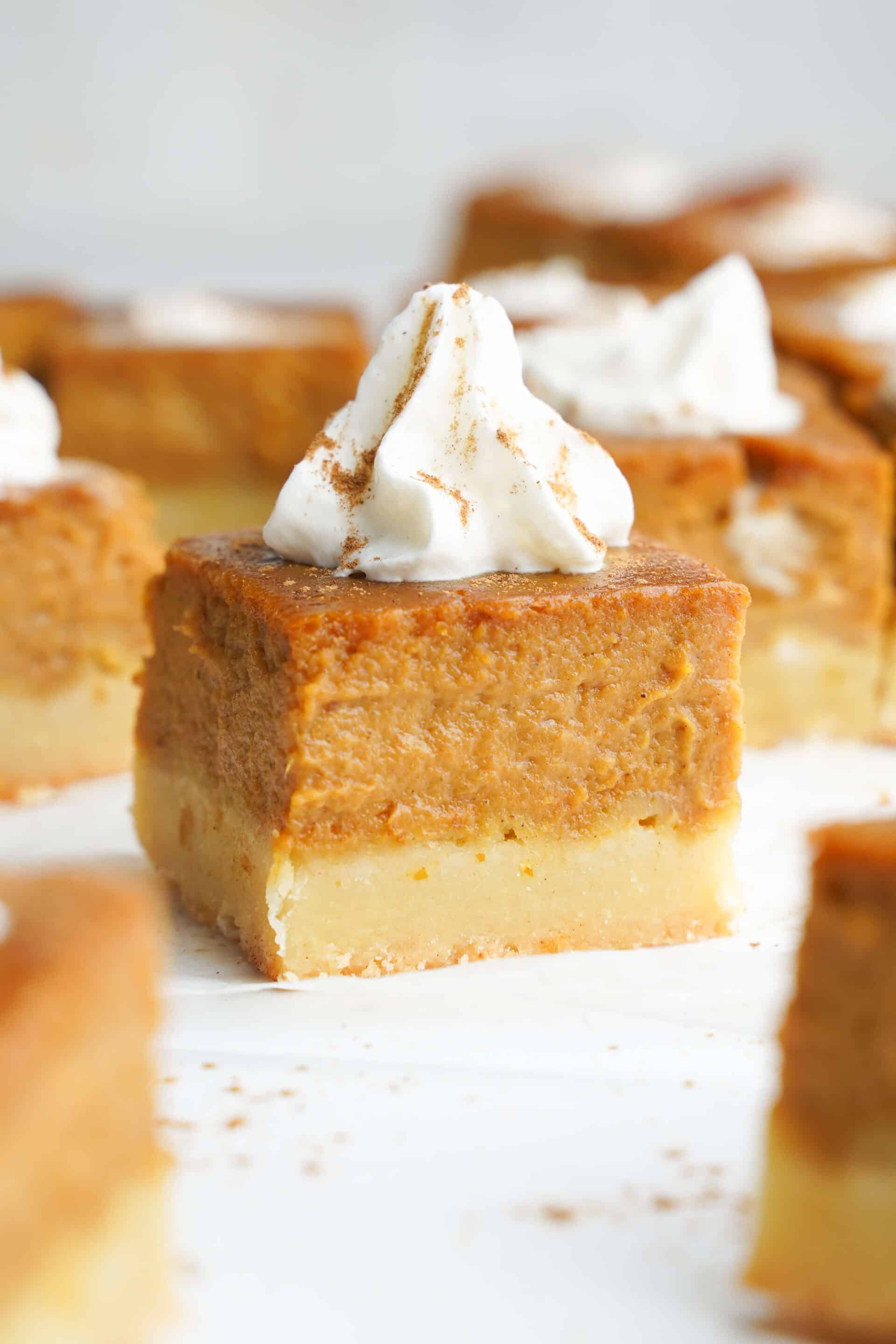 A close-up of a pumpkin pie bar with a thick pumpkin filling on a shortbread crust, topped with whipped cream and a dusting of cinnamon.