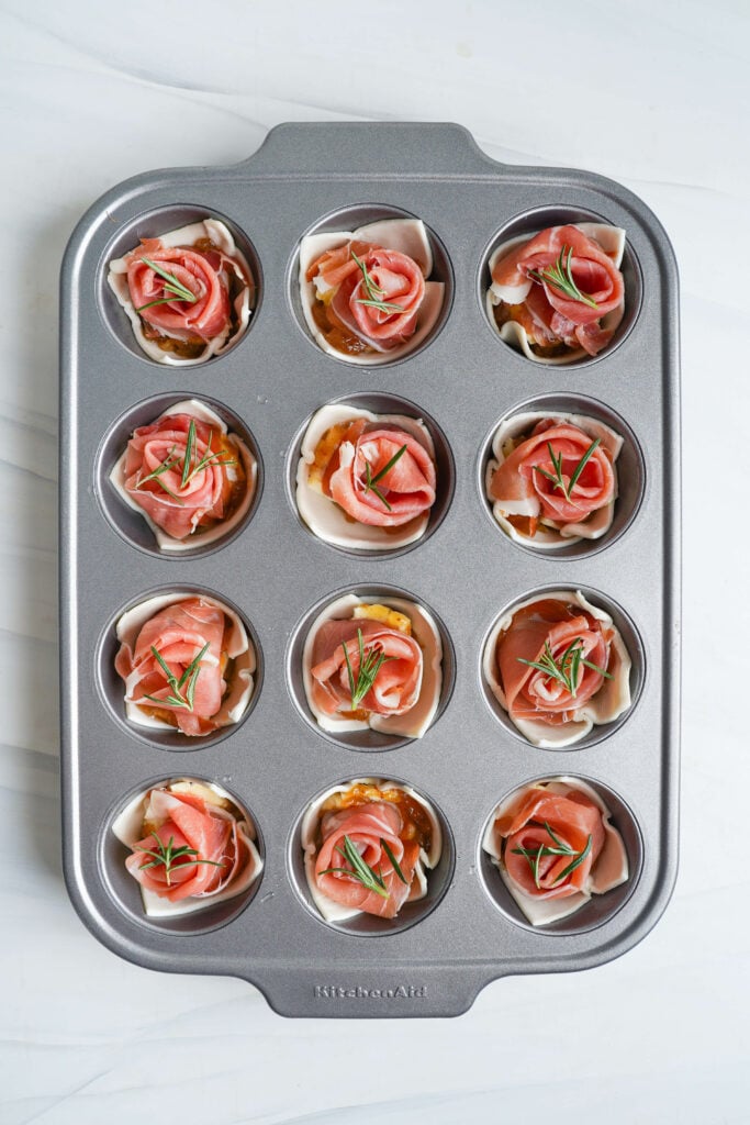 Unbaked brie and puff pastry bites topped with prosciutto and fresh rosemary in a muffin tin before baking.