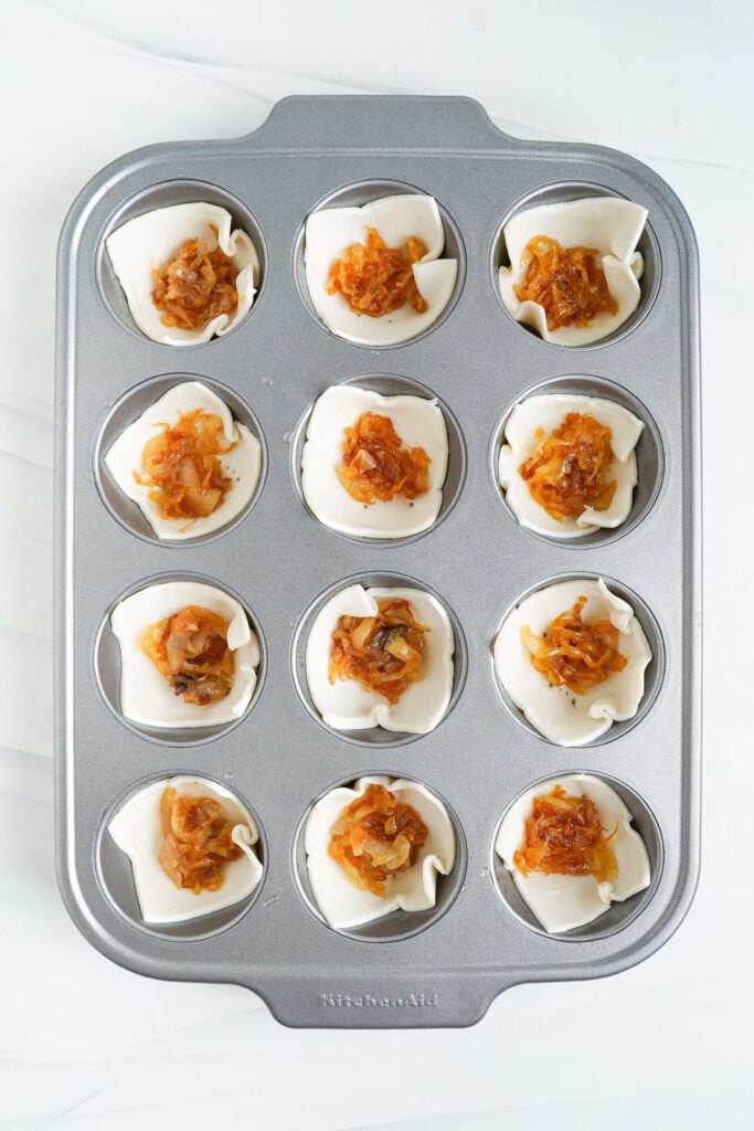 Muffin tin filled with puff pastry squares topped with a spoonful of caramelized onions as the first filling layer.