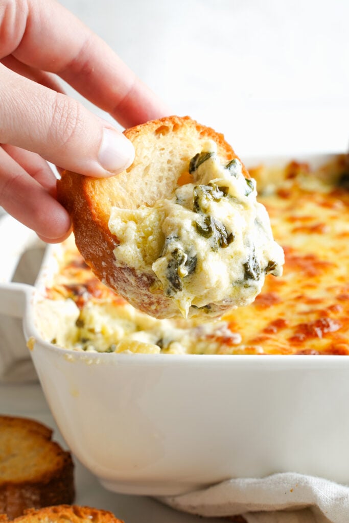 Toasted baguette being dipped into hot, creamy spinach artichoke dip straight from the baking dish.