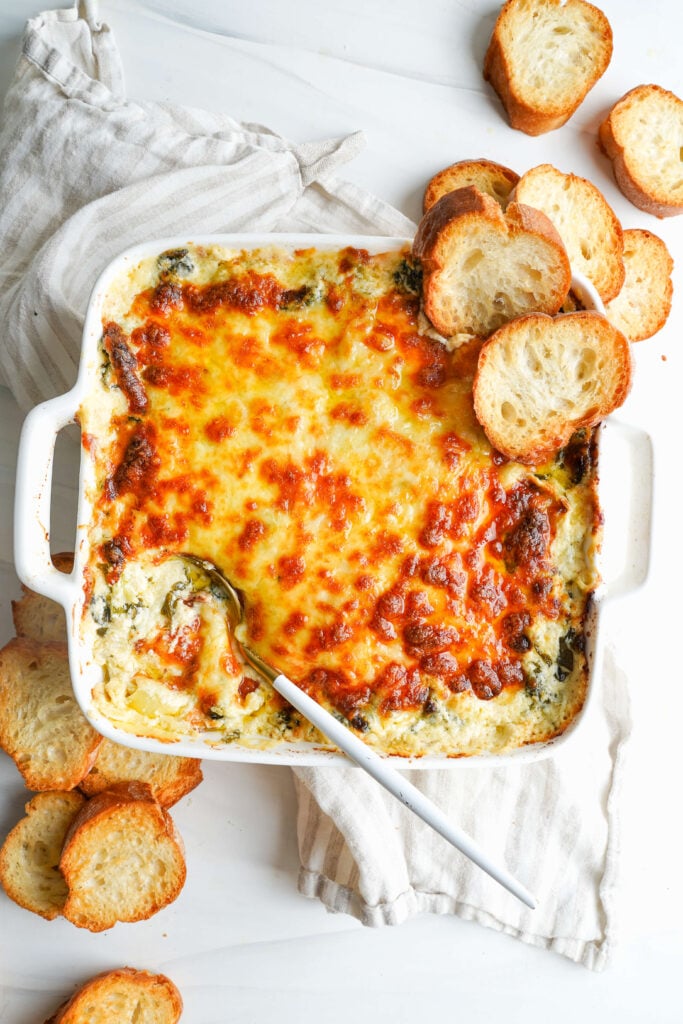 Hot spinach artichoke dip in a baking dish with toasted baguette slices for serving.