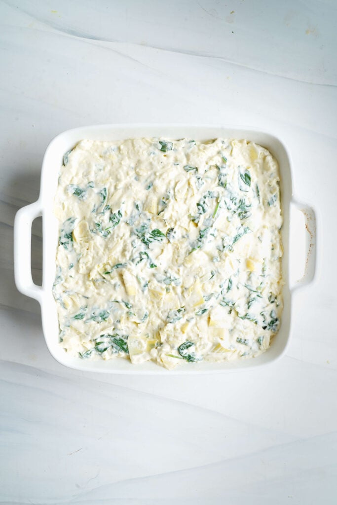 Unbaked spinach artichoke dip spread evenly in a white baking dish, ready to be topped with cheese.
