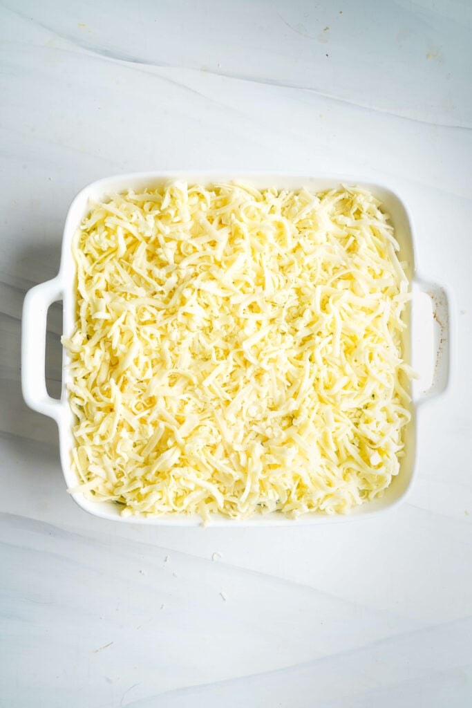 Baking dish filled with spinach dip topped with a thick layer of shredded white cheddar before baking.