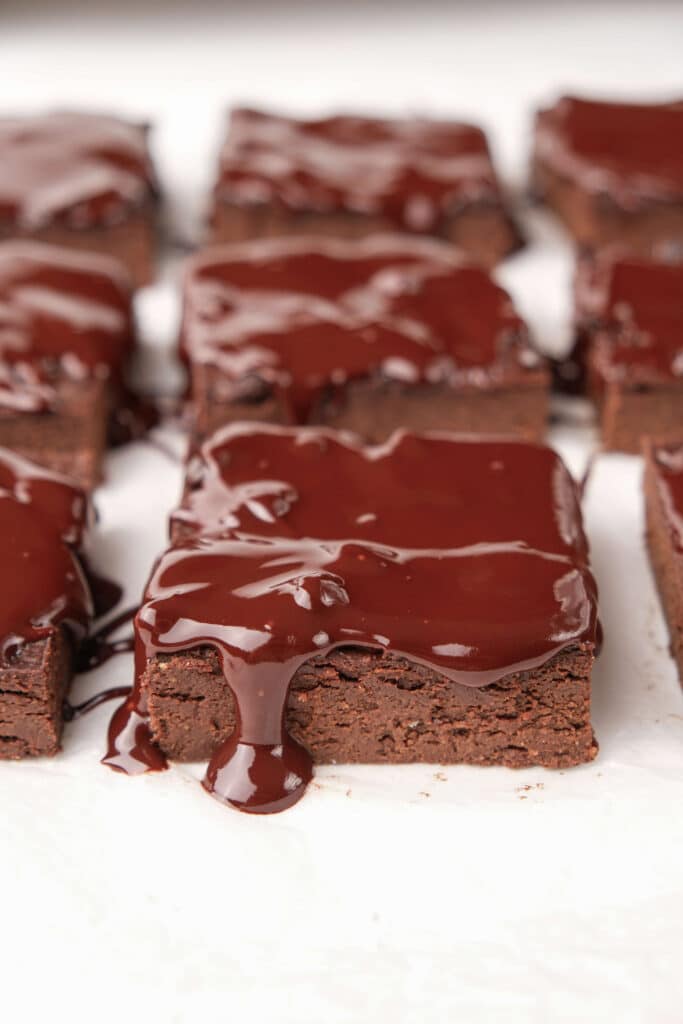 Square avocado brownies topped with smooth chocolate fudge frosting, showing a thick chocolate drip and a dense interior.