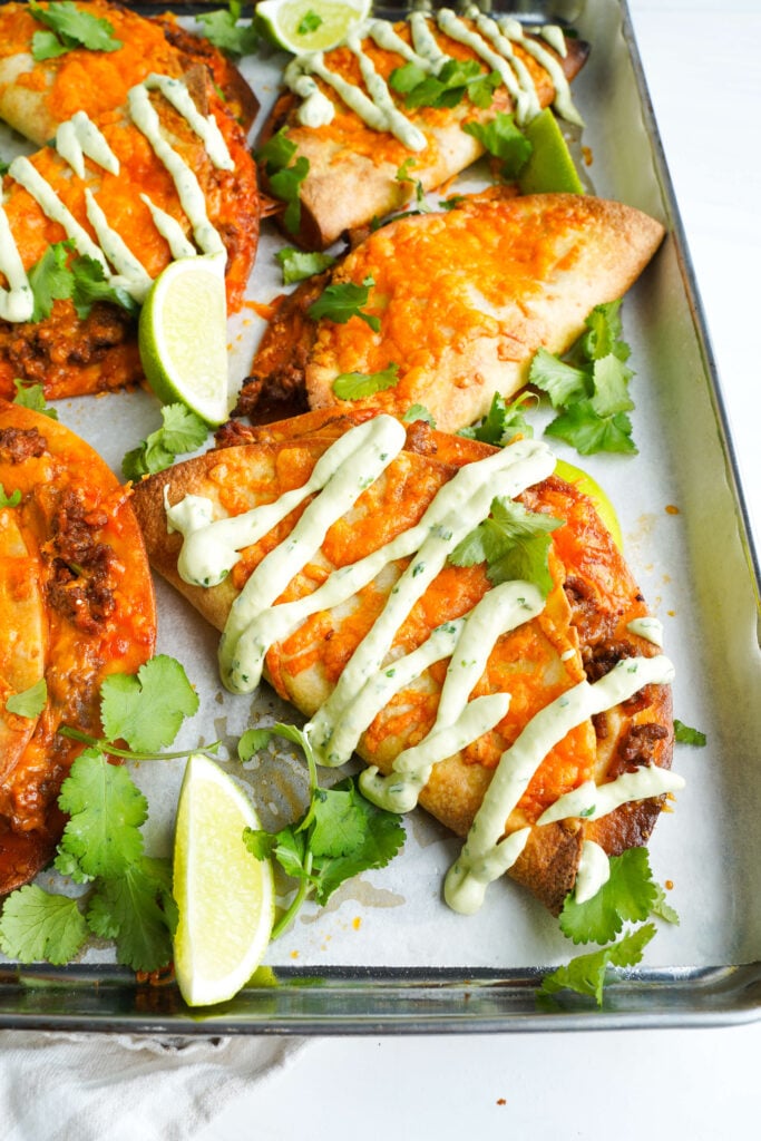 Crispy baked tacos filled with seasoned ground beef and melted cheddar cheese, drizzled with avocado lime crema and garnished with fresh cilantro and lime wedges on a sheet pan.