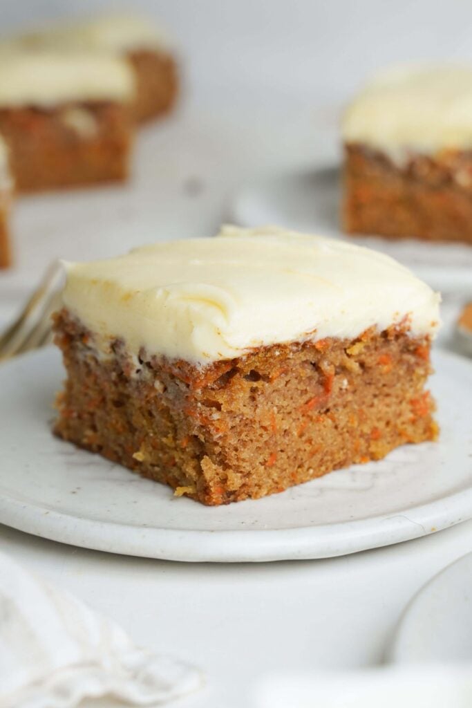 A slice of carrot cake with cream cheese frosting on a white plate with additional pieces in the background