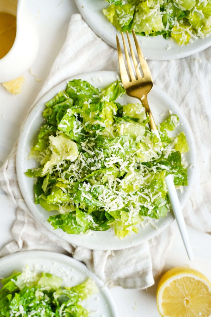 Three plates of lemon parmesan salad topped with finely grated Parmesan cheese on a white cloth with a lemon half nearby