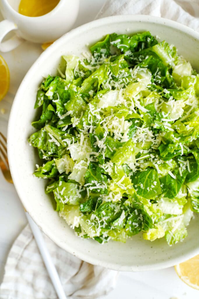 Bowl of chopped romaine lettuce topped with finely grated Parmesan cheese, with a lemon half and white pitcher nearby