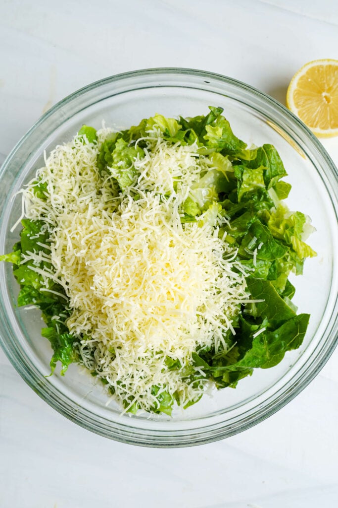 Chopped romaine lettuce in a glass bowl topped with freshly grated Parmesan cheese and a lemon half nearby