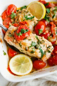 Baked halibut with cherry tomatoes, garlic, parsley, and lemon in a white baking dish with a spoon lifting a piece of fish