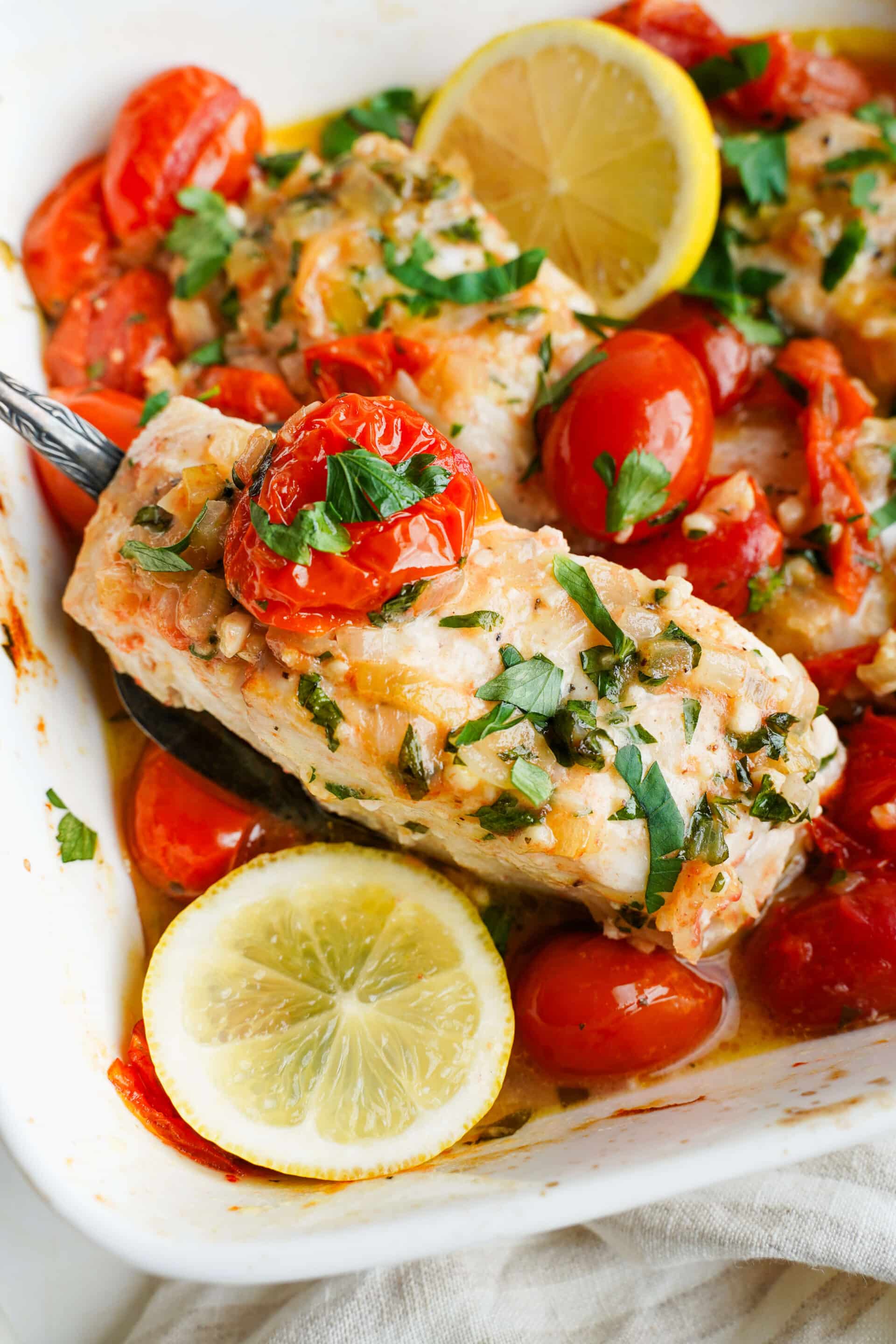 Baked halibut with cherry tomatoes, garlic, parsley, and lemon in a white baking dish with a spoon lifting a piece of fish