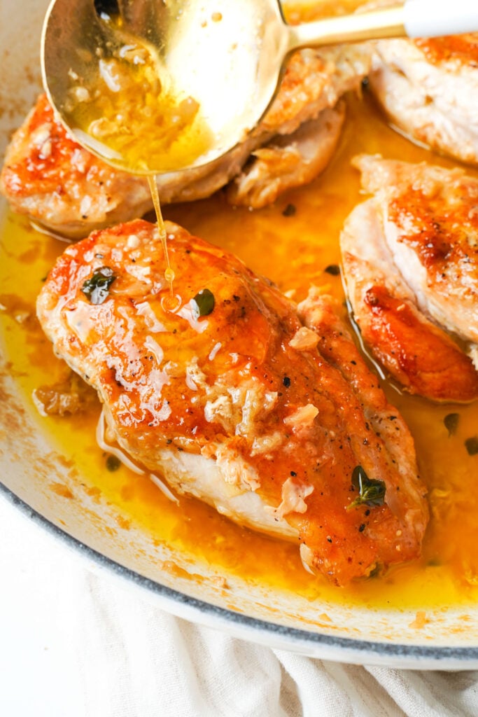 Golden chicken breasts in a skillet with a buttery pan sauce being spooned over the top.