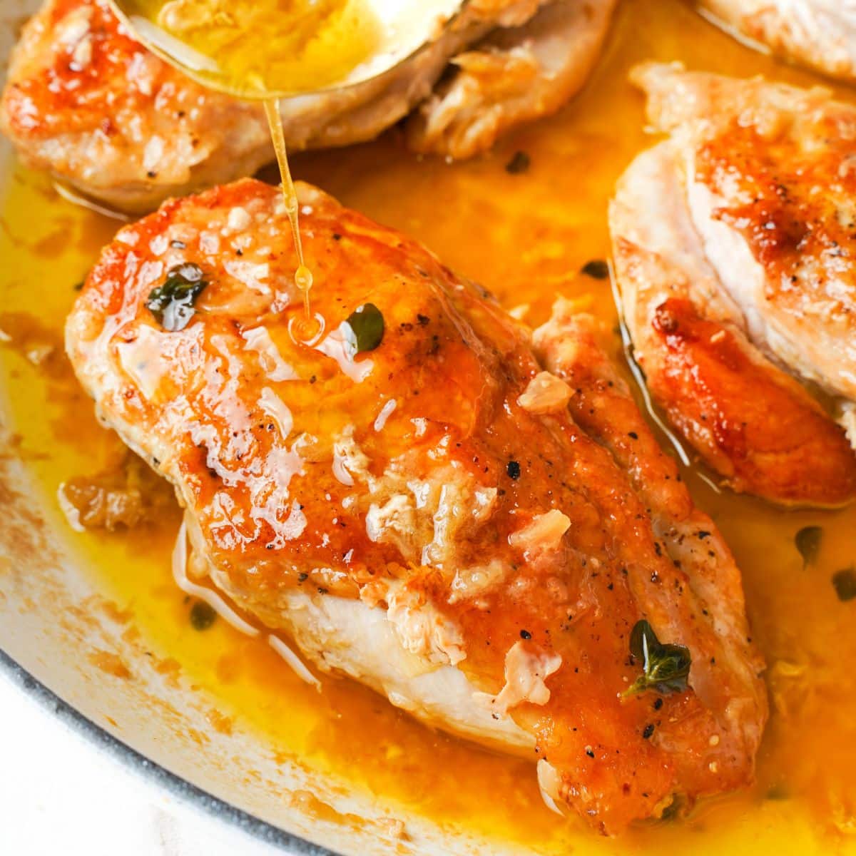 Golden chicken breasts in a skillet with glossy pan sauce, fresh thyme, and sauce being spooned over the top.