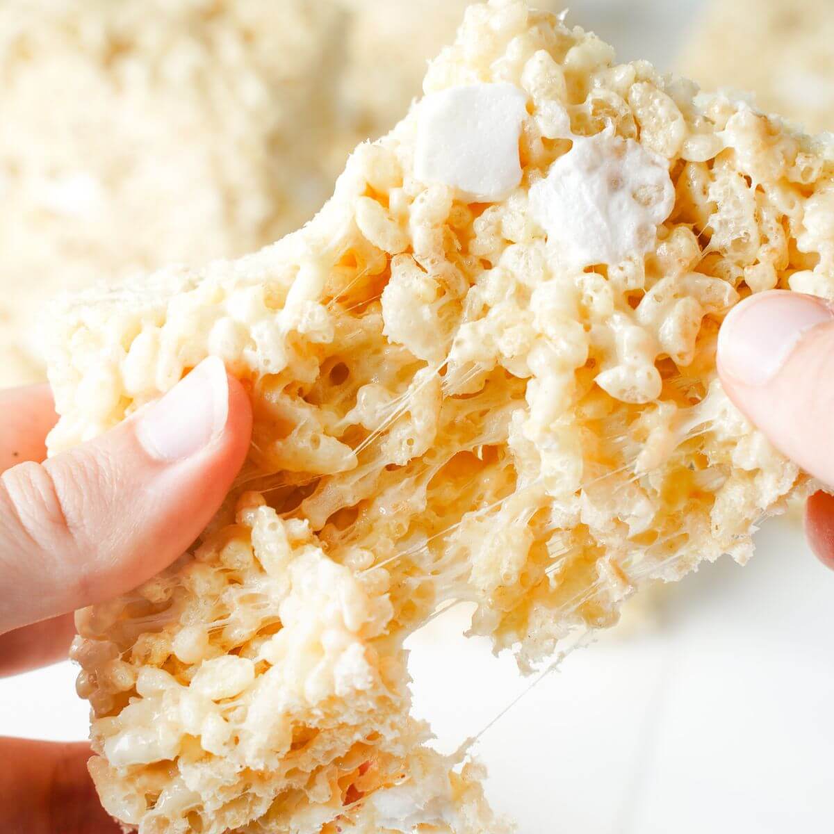 Hands pulling apart a gooey Rice Krispie treat with melted marshmallow stretching between the cereal and soft marshmallow pockets visible throughout.
