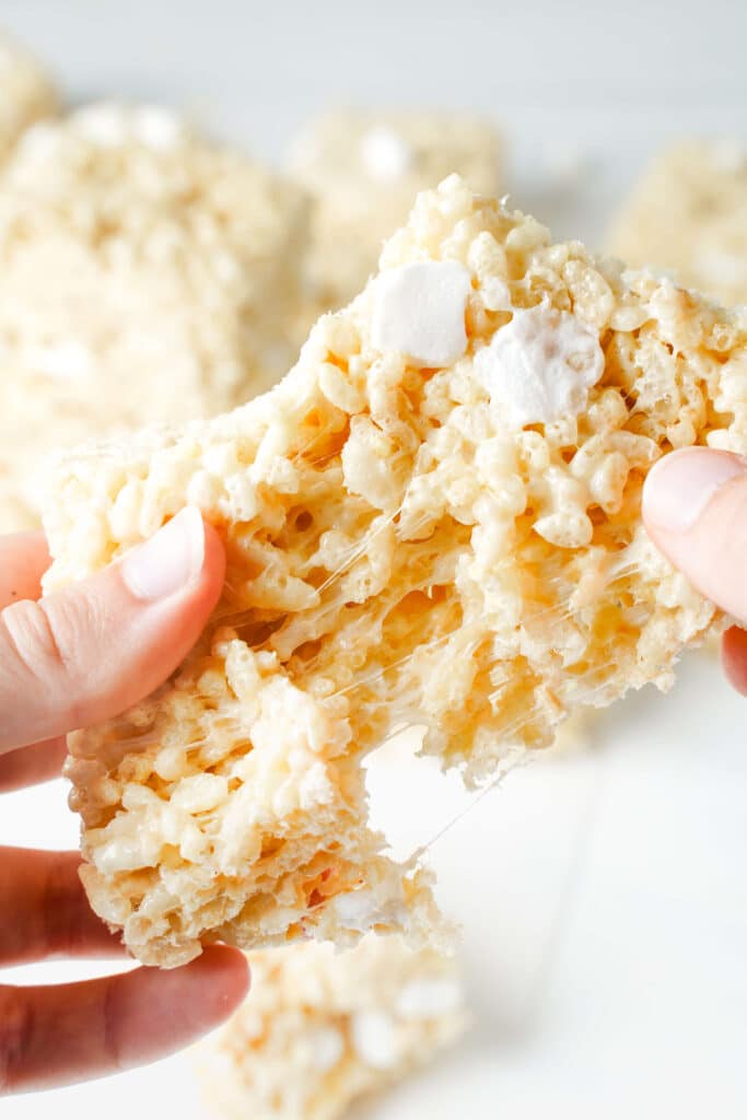 Hands pulling apart a gooey Rice Krispie treat with melted marshmallow stretching between the cereal and soft marshmallow pockets visible throughout.