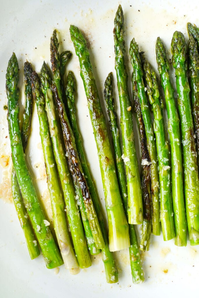 Sautéed asparagus spears with olive oil, minced garlic, black pepper, and lightly charred edges on a white plate.