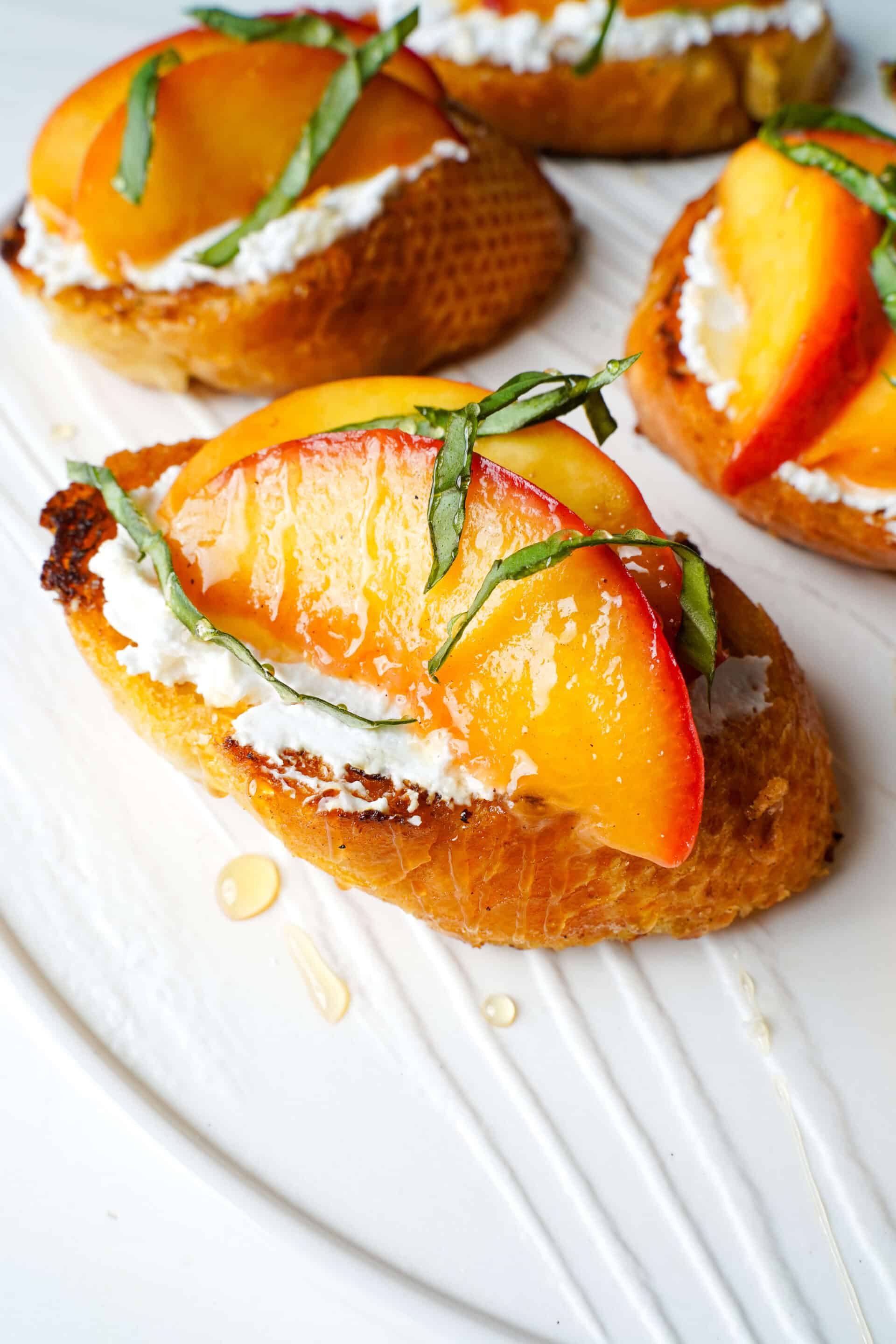 Close up shot of easy peach bruschetta featuring golden pan-fried baguette slices topped with creamy goat cheese, warm caramelized peaches, honey drizzle, and fresh basil.